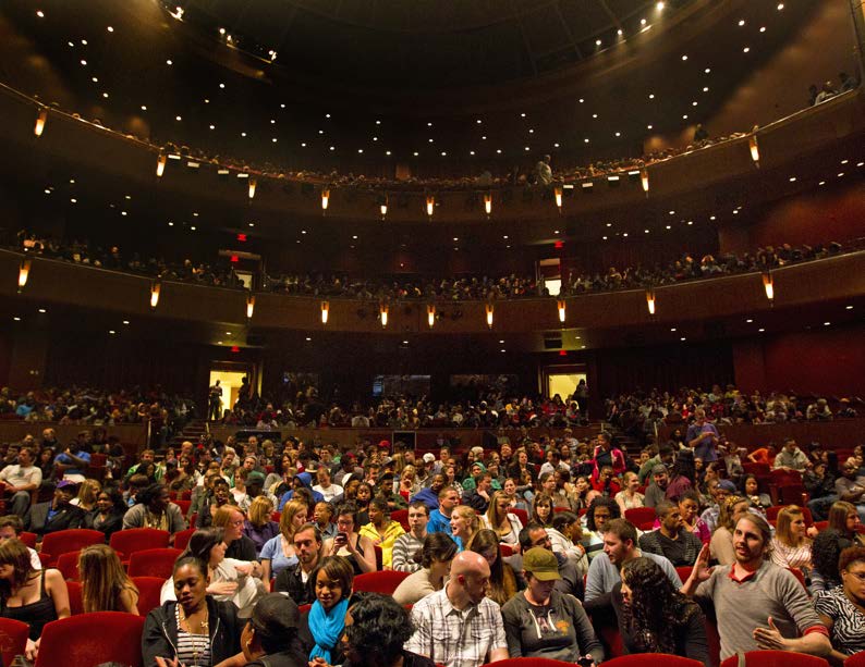 Crowded theater auditorium with audience seated across multiple balcony levels.